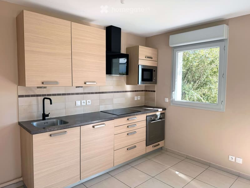 Modern kitchen with light wood cabinets, stainless steel appliances, and a large window overlooking greenery.