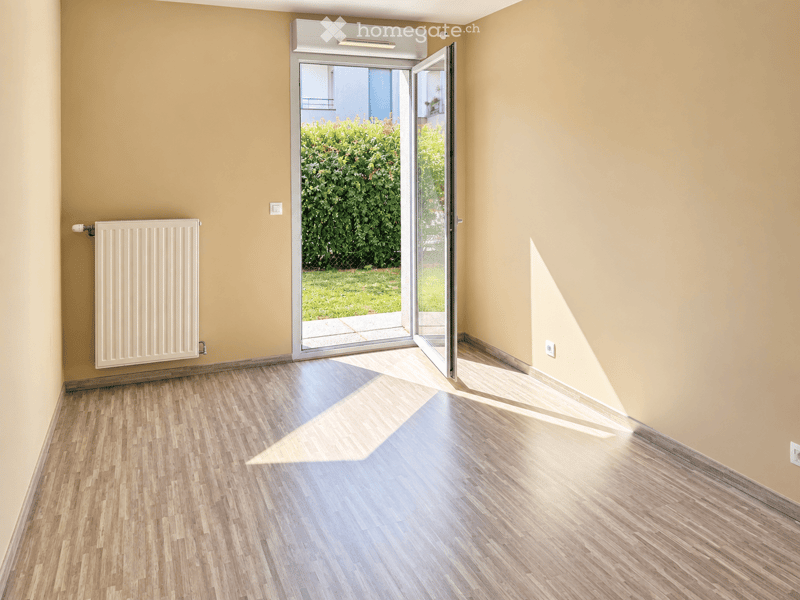 Empty room with light yellow walls, wooden flooring, and a white radiator on one wall. A large sliding glass door opens to a green garden area.