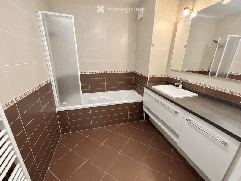 Modern bathroom with brown tiled walls and floor, white bathtub, white sink with mirror, and white cabinet.