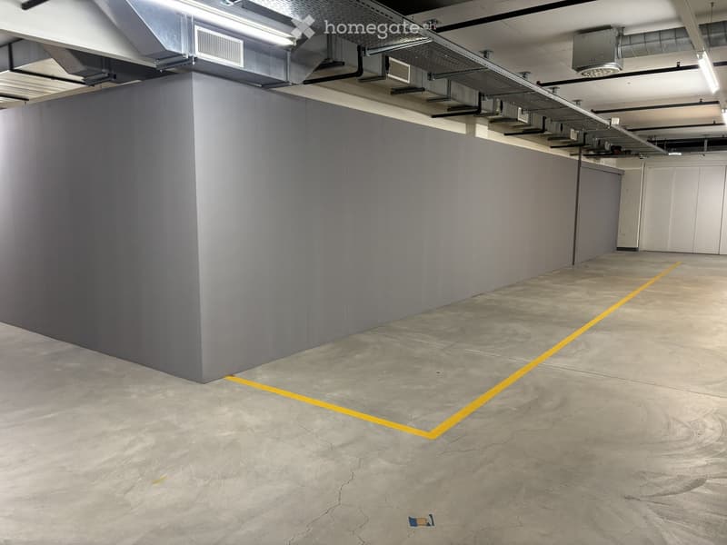 Empty underground parking space with grey concrete walls, concrete floor, yellow parking lines, and exposed ceiling pipes and lighting.