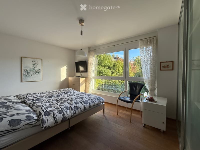 A modern bedroom with a bed, nightstand, chair, lamp, television, and picture frame. Large windows offer a view of greenery outside.