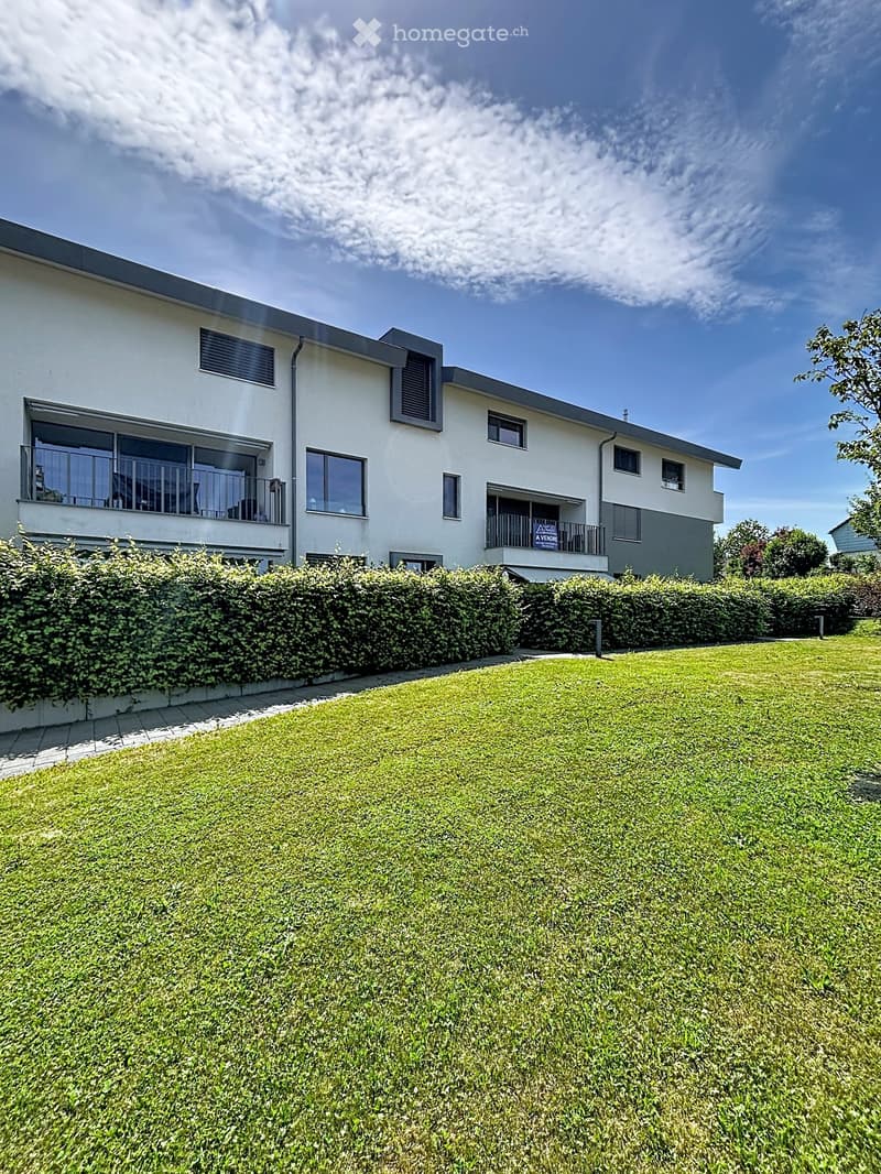 Modern two-story house with white exterior, large windows, and balconies, surrounded by a well-maintained lawn and hedges.