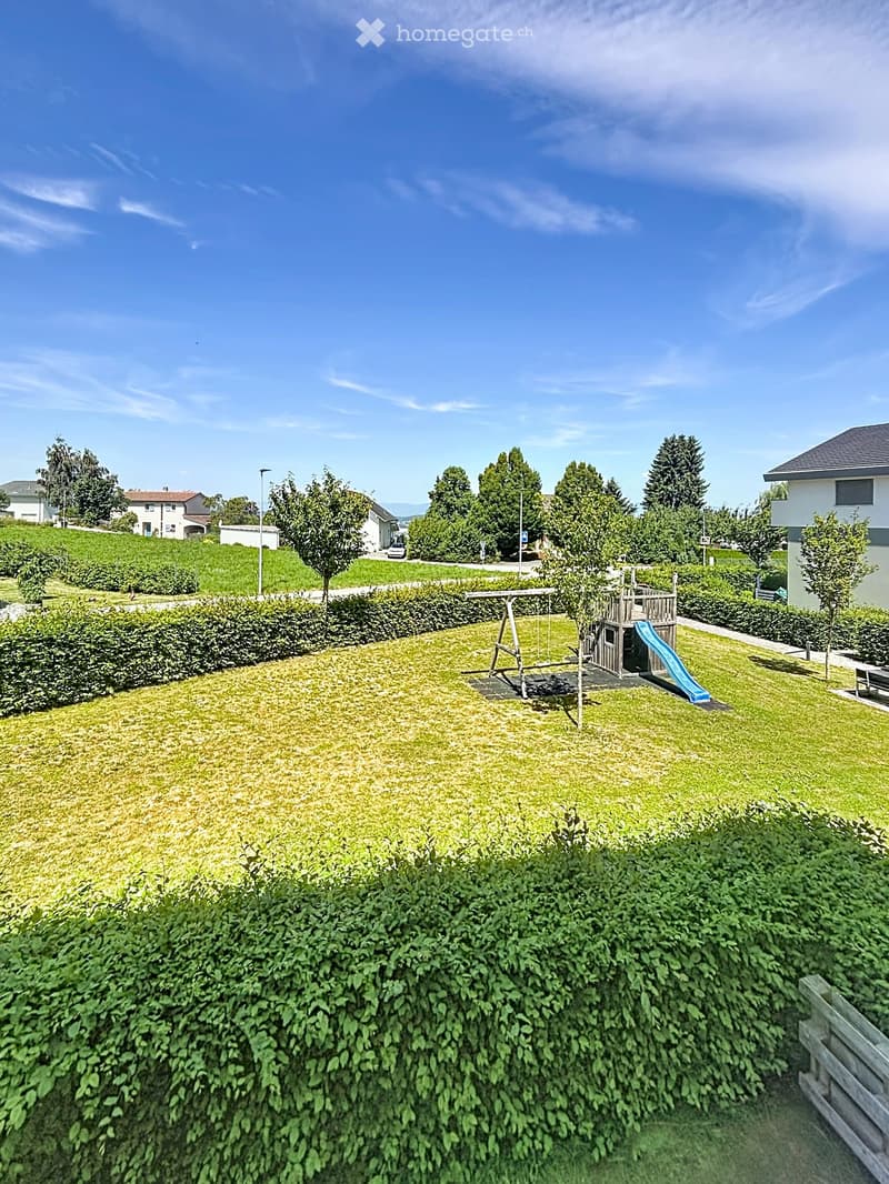 A well-maintained garden with a swing set, slide, and surrounding greenery, bordered by hedges and trees.