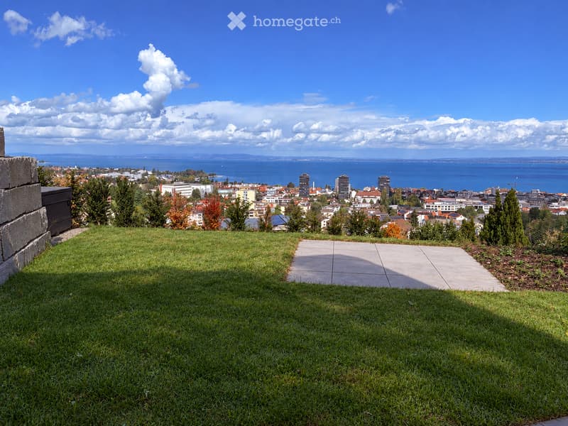 A grassy area with a paved pathway leading towards a cityscape view, with trees and buildings visible in the distance under a partly cloudy sky.