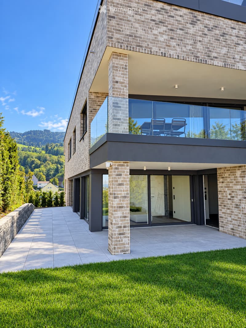 Modern two-story house with brick facade, large glass windows, and a spacious balcony overlooking a scenic valley.