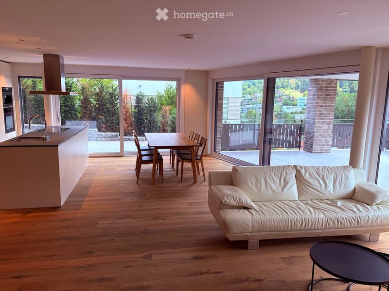 Open-plan living and dining area with a white leather sofa, coffee table, dining table with chairs, and a kitchen island with a faucet. Large windows offer a scenic view of greenery and a balcony.