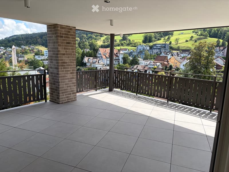 Balcony with tiled floor, dark wooden railing, and scenic view of a town and green hills.