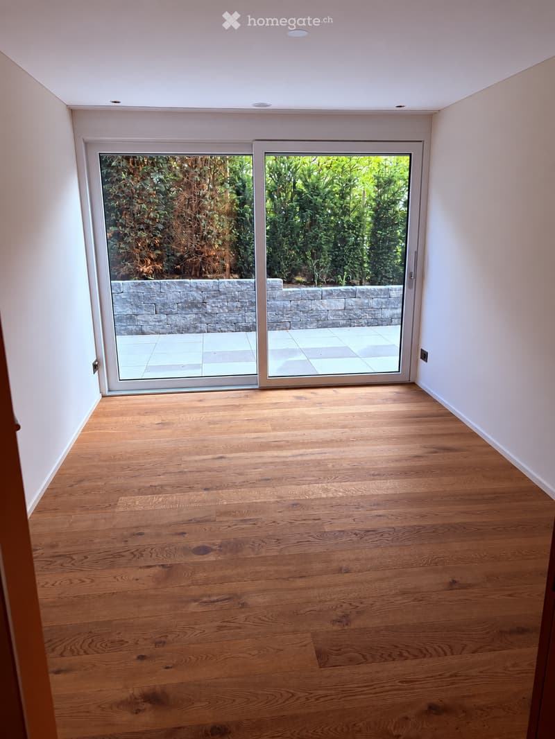 Empty room with wooden flooring, white walls, and large sliding glass doors leading to an outdoor area with a stone wall and greenery.