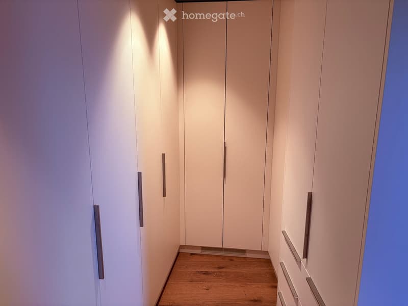 A narrow hallway with light-colored, modern built-in wardrobes and a wooden floor. The wardrobes have sleek, metallic handles and are illuminated by soft, ambient lighting.