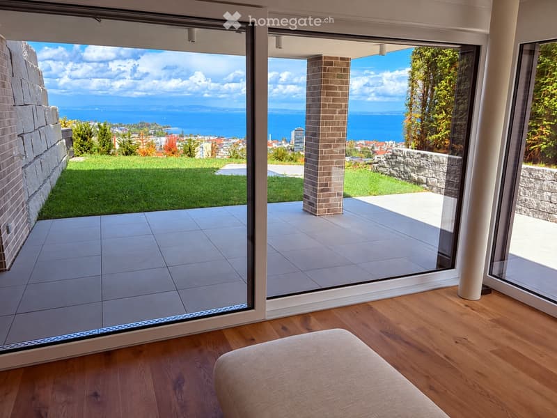 Modern interior with wooden flooring, large glass sliding doors opening to a balcony with scenic ocean view, and a light-colored sofa visible.