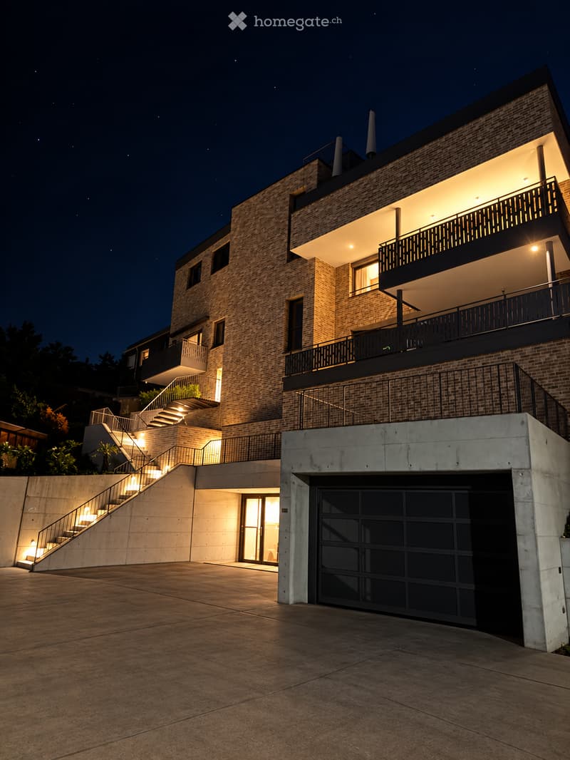 Modern two-story house with brick facade, illuminated at night, featuring a raised ground floor, garage, and multiple balconies.