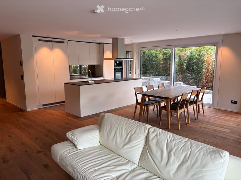 Modern open-plan living and dining area with a white sofa, wooden dining table with chairs, and a kitchen island with a sink and oven. Large windows provide a view of the garden.