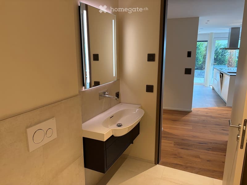 Modern bathroom with a white sink, dark cabinet, and large mirror. The room has light-colored tiles and a wooden floor visible in the adjacent space.