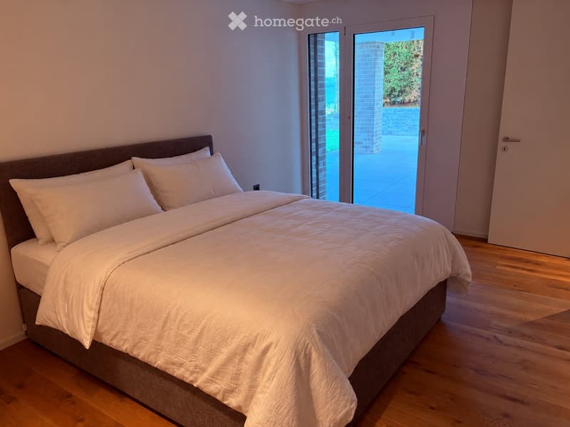 Modern bedroom with a large bed, white bedding, and wooden flooring. Large sliding glass doors lead to an outdoor area.