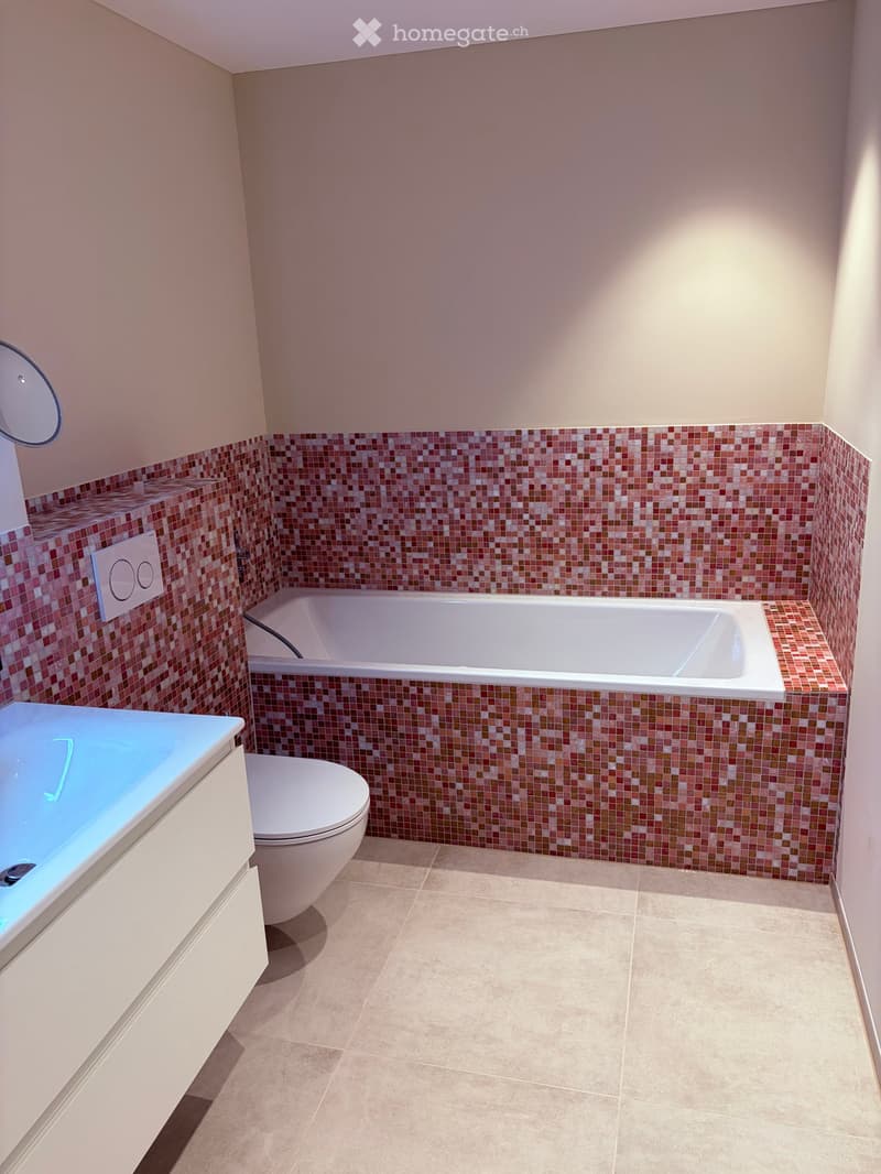 Modern bathroom with a bathtub, toilet, sink, and mirror. The walls are tiled with a mosaic pattern in red and beige, and the floor is tiled in beige.