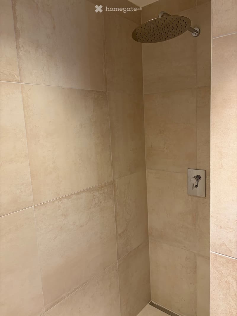 Modern bathroom corner with beige tiled walls, a round showerhead mounted on the wall, and a chrome control valve.