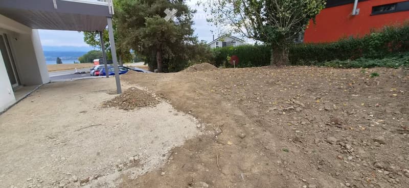 An outdoor area with loose soil and rocks, partially covered with grass, with a modern house and parked cars visible in the background.