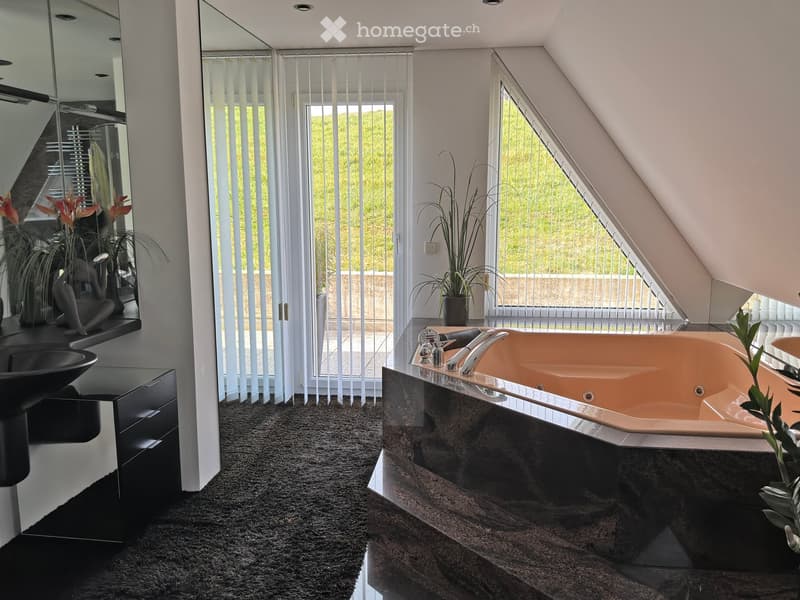 Modern bathroom with a large bathtub, sleek black sink, and large windows with blinds. Features include a dark carpeted floor, a mirror, and decorative plants.