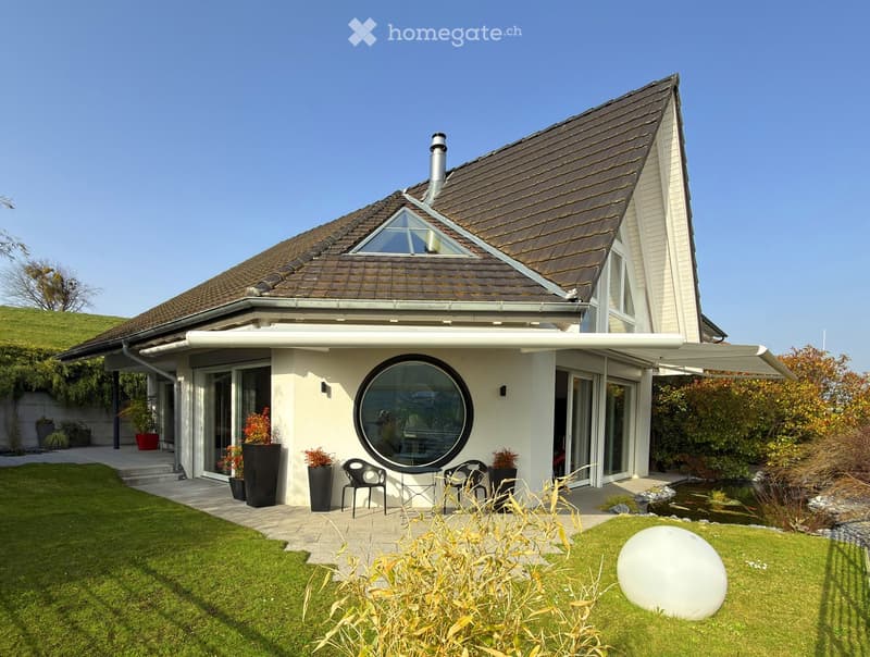 Modern two-story house with a brown tiled roof, large windows, and a spacious garden featuring a pond, patio seating, and decorative plants.