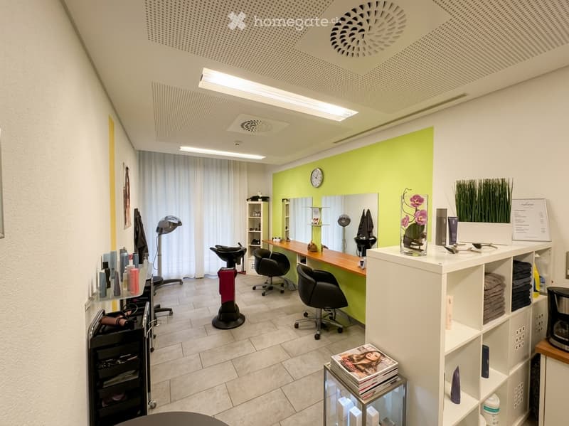 Modern, well-lit salon with a bright green accent wall, large mirror, multiple chairs, a central table, shelves with products, and decorative vases.