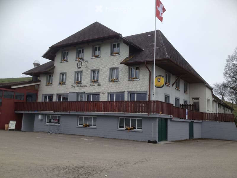 Two-story building with white brick facade, multiple windows, and a balcony with wooden railing. A Swiss flag is displayed on a flagpole, and there is signage for a restaurant named "Berg Restaurant Ahorn Alp." The building has a gray lower level and a br