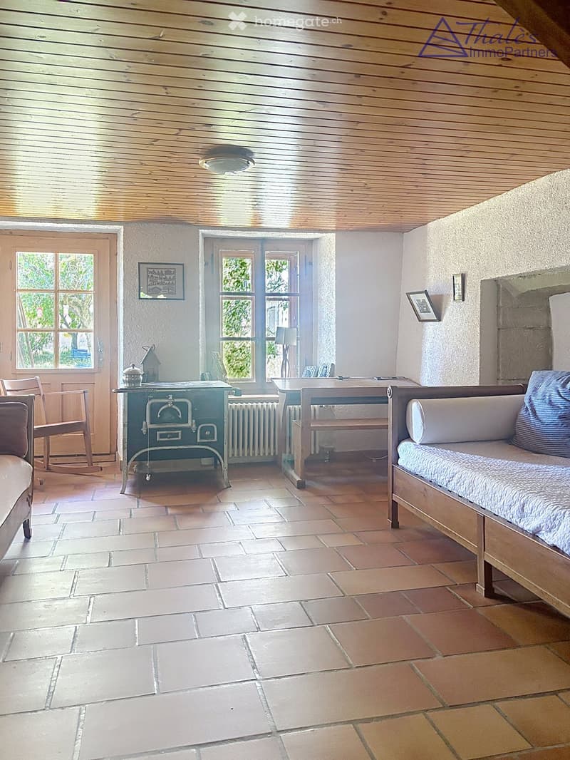Cozy room with tiled floor, wooden ceiling, and large windows. Features a sofa, chairs, a small table, and a radiator.