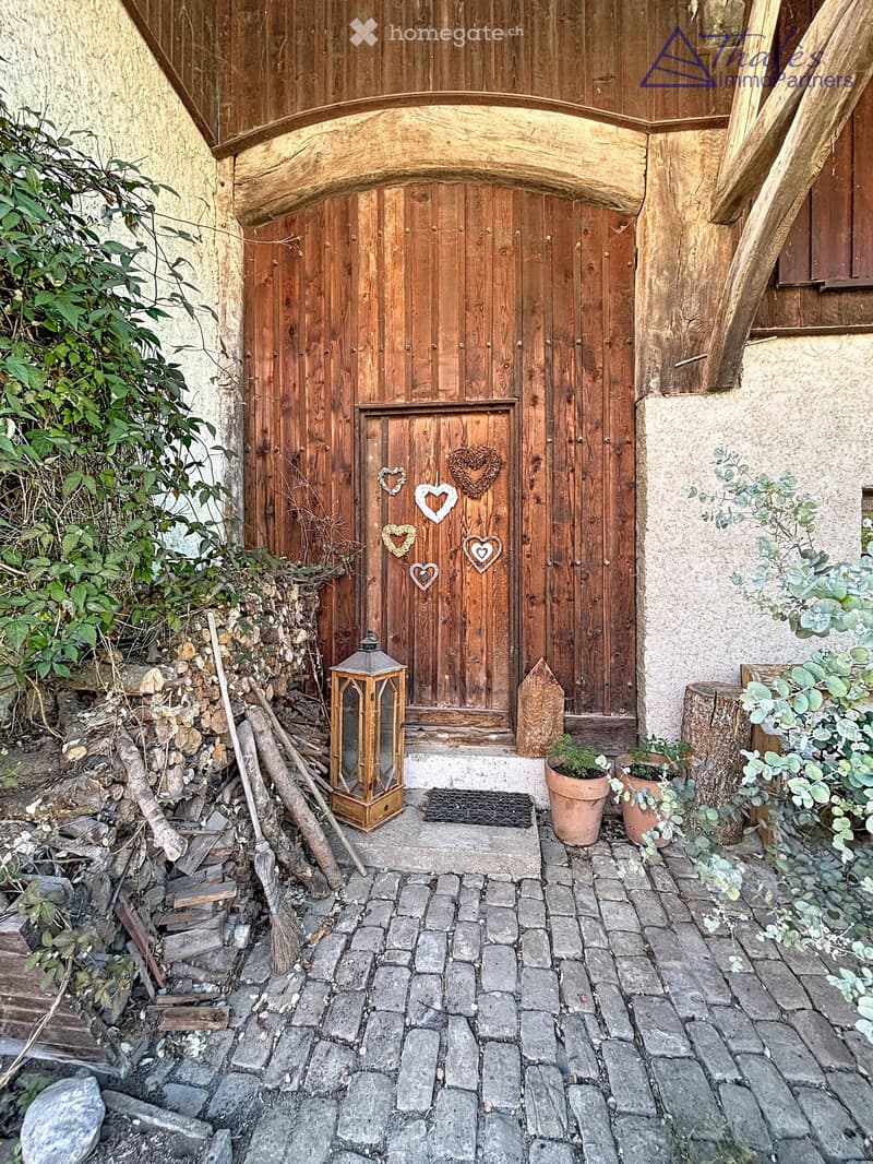 Entrance to a rustic building with a large wooden door adorned with heart-shaped decorations, a lantern, potted plants, and a cobblestone pathway.