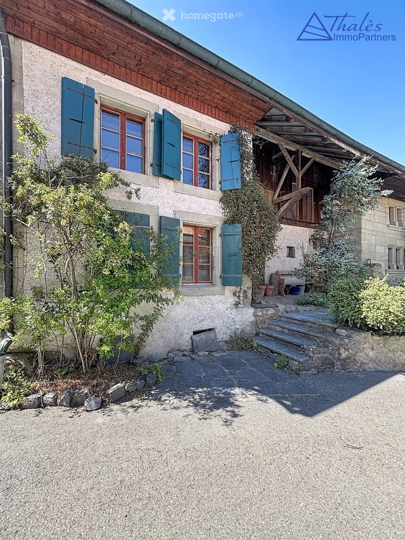 A rustic stone house with wooden shutters and a covered wooden porch, surrounded by greenery and a gravel driveway.