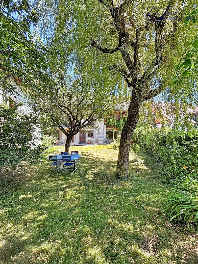 Lush garden with a large tree, a small table and chairs, and a house in the background.