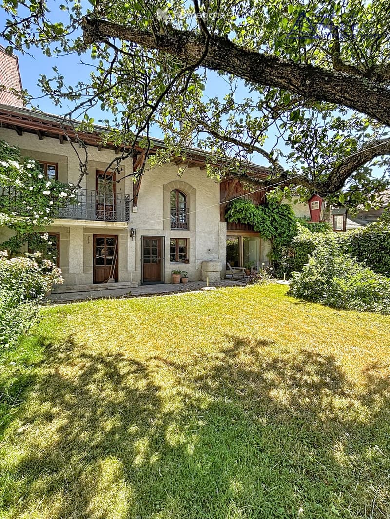A two-story house with beige stone walls, wooden doors and windows, a balcony with metal railings, and a well-maintained lawn.