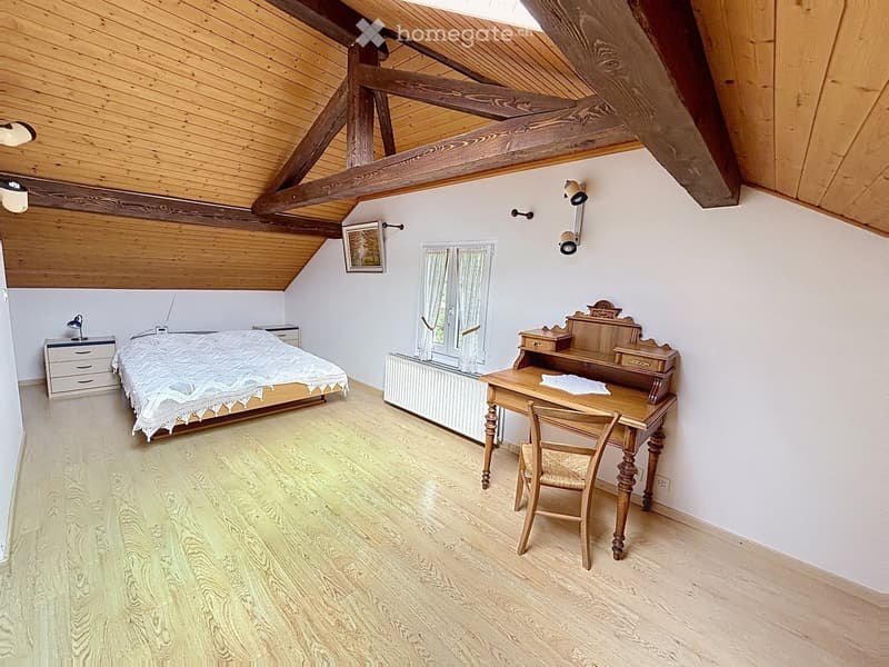 Cozy attic bedroom with wooden slanted ceilings, a double bed with white bedding, a wooden desk with a chair, a nightstand, and a window with white curtains.