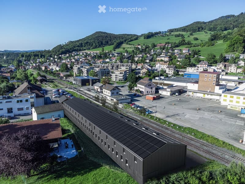 Aerial view of a town with a mix of residential and commercial buildings, surrounded by green hills and a clear blue sky.
