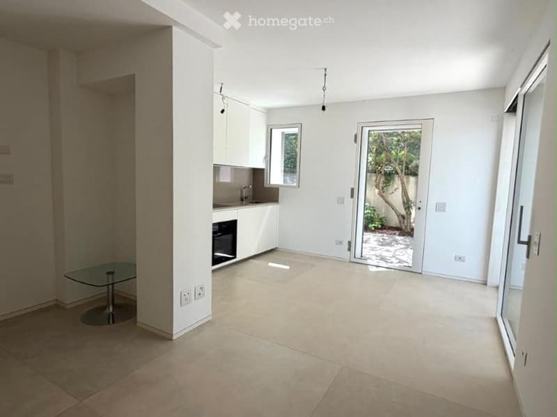 Open-plan unfurnished room with tiled flooring, white walls, built-in kitchen cabinets, and large sliding glass doors leading to a balcony area.
