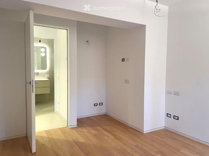 Empty room with light wooden flooring and white walls, featuring an open doorway leading to a small bathroom with a sink and mirror.