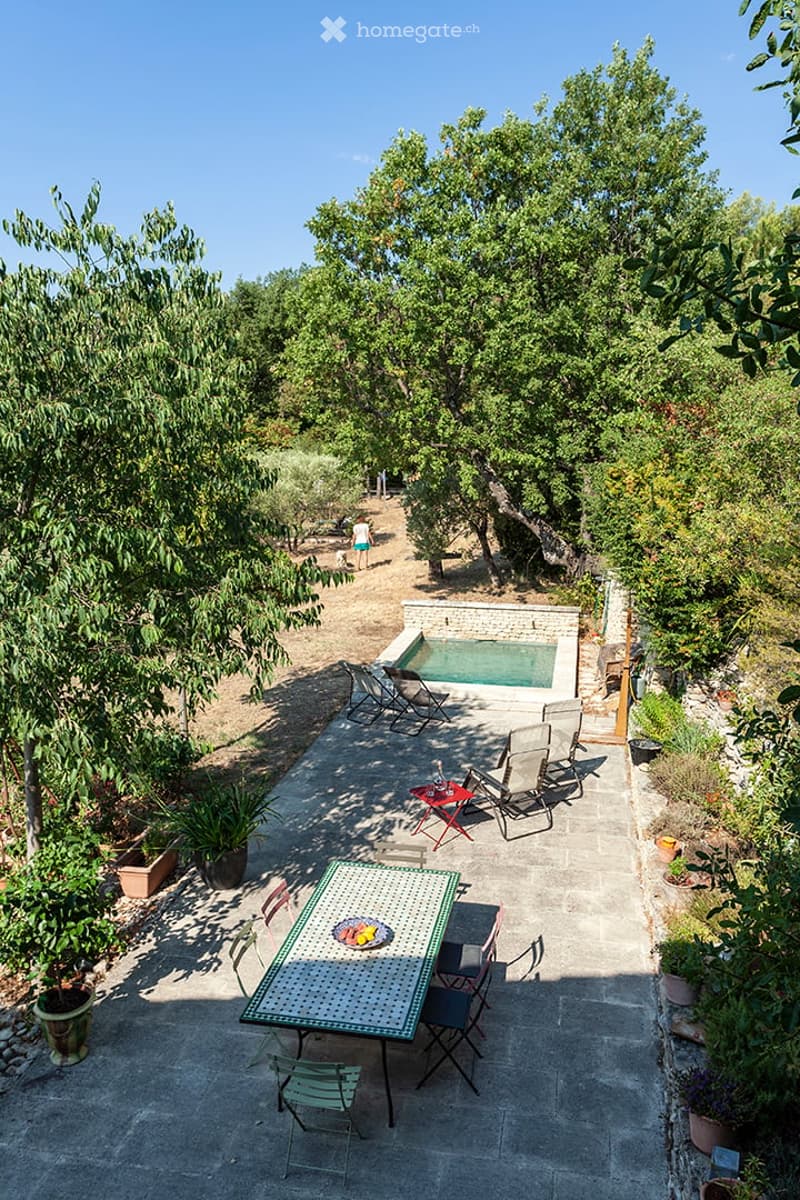Outdoor patio with a dining table, chairs, and a swimming pool surrounded by trees and greenery.