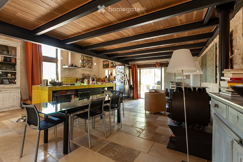 Open-plan living and dining area with exposed wooden beams, a glass dining table with chairs, a sofa, a floor lamp, and a kitchen area with a counter, refrigerator, and shelves.