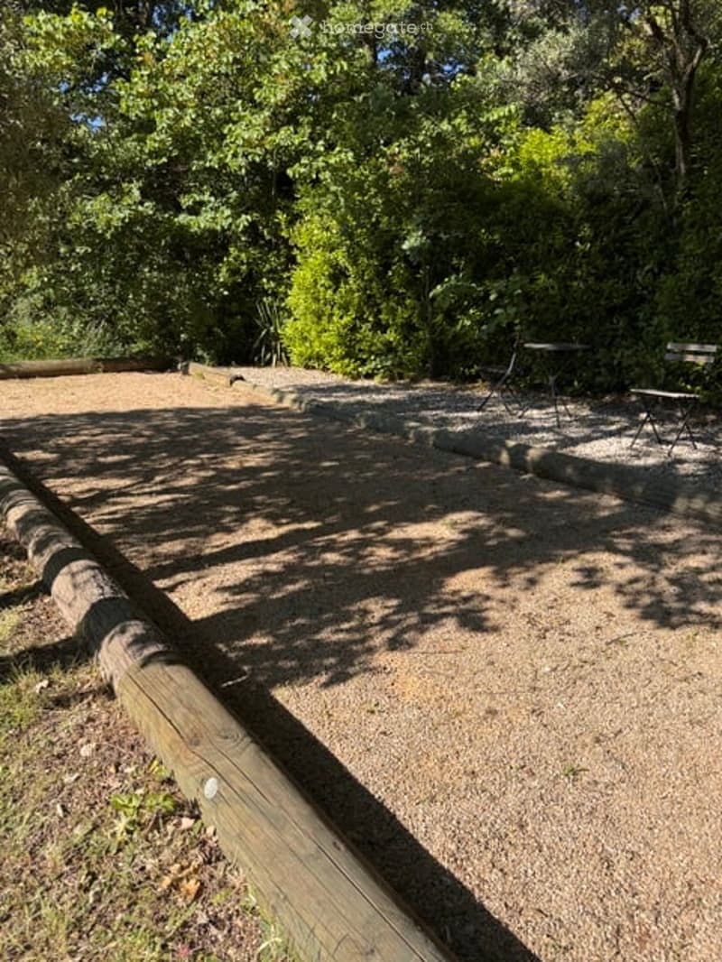 Outdoor area with gravel ground, wooden bench, small table and chairs, surrounded by trees and greenery.