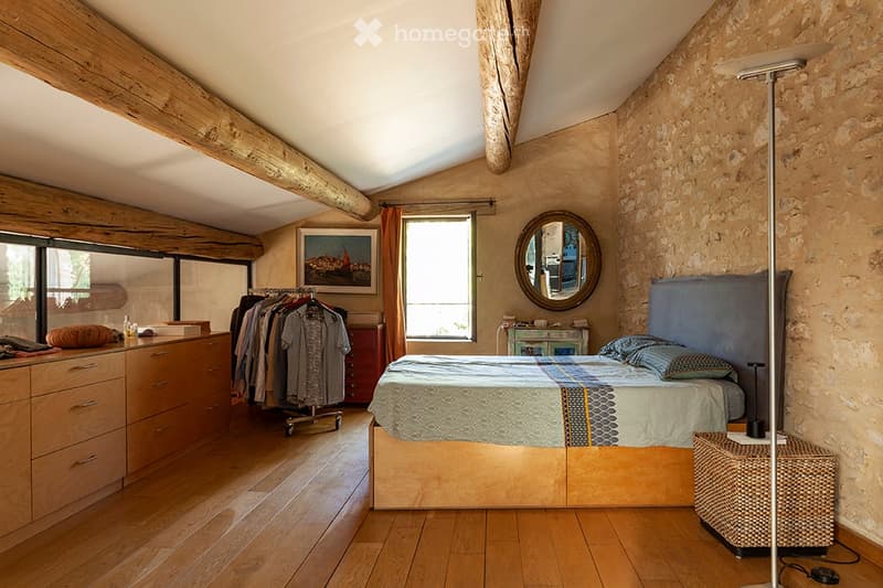 Cozy bedroom with exposed wooden beams, stone walls, and wooden flooring. Features a large bed with a patterned duvet, a nightstand, a round mirror, a painting, a clothing rack, and a wicker basket.