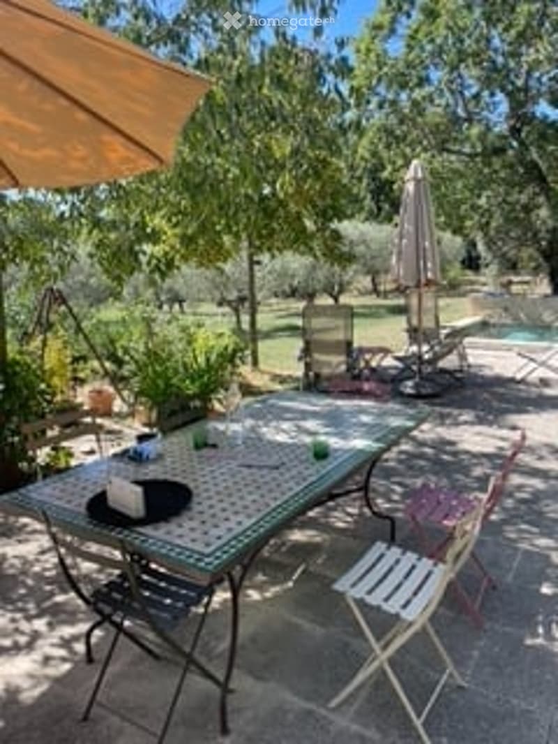 Outdoor patio area with a table, chairs, umbrella, and a swimming pool in the background.