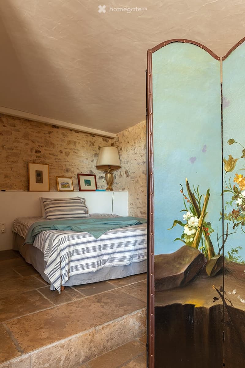 Cozy bedroom with stone walls, a bed with striped bedding, a table lamp, and a decorative mirror with floral artwork.