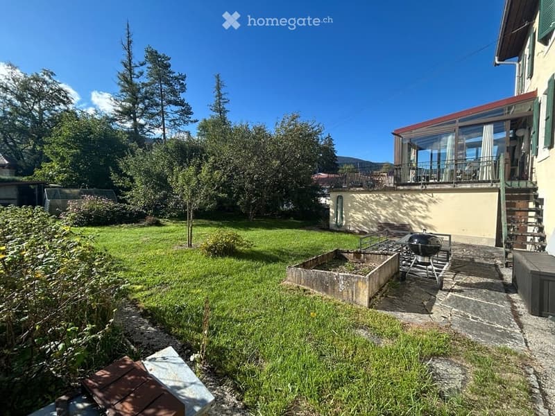Well-maintained garden with a lawn, trees, a raised garden bed, and a barbecue. The house features a balcony with large windows and a staircase leading to the garden.