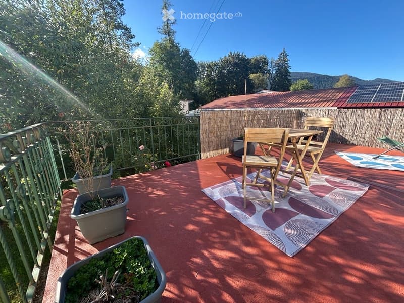 Balcony with wooden folding chairs and table, potted plants, decorative rug, and a view of greenery and solar panels on the roof.