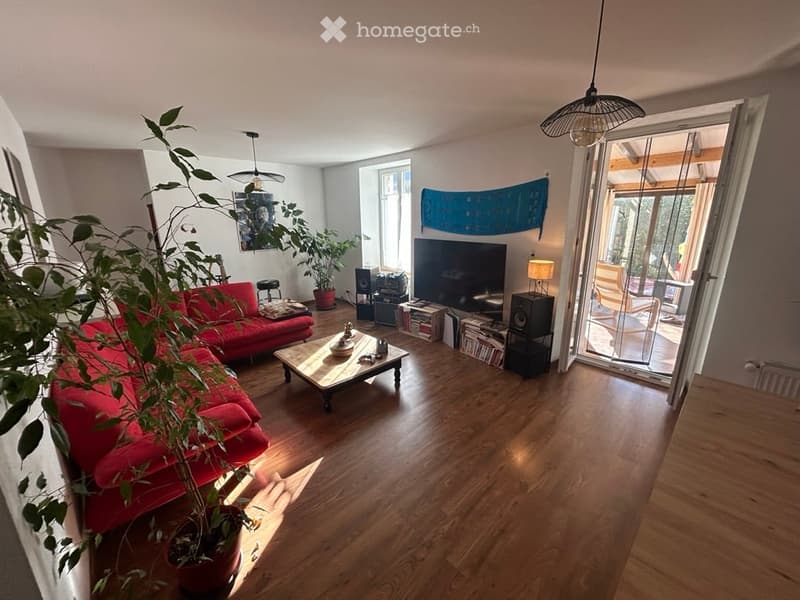 Bright living room with wooden flooring, red sofa, coffee table, potted plants, TV, speakers, and a view to a balcony or terrace.