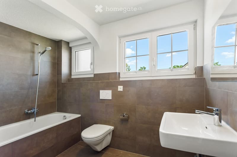 Modern bathroom with a bathtub, toilet, sink, and showerhead. Brown tiled walls and floor, large windows letting in natural light.
