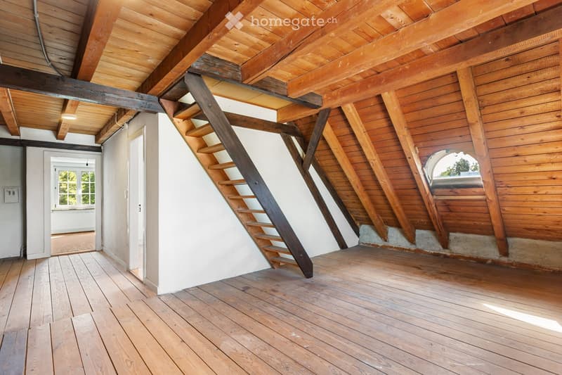 An empty attic space with wooden beams, exposed wooden ceiling, wooden floor, and a narrow wooden staircase leading upwards.