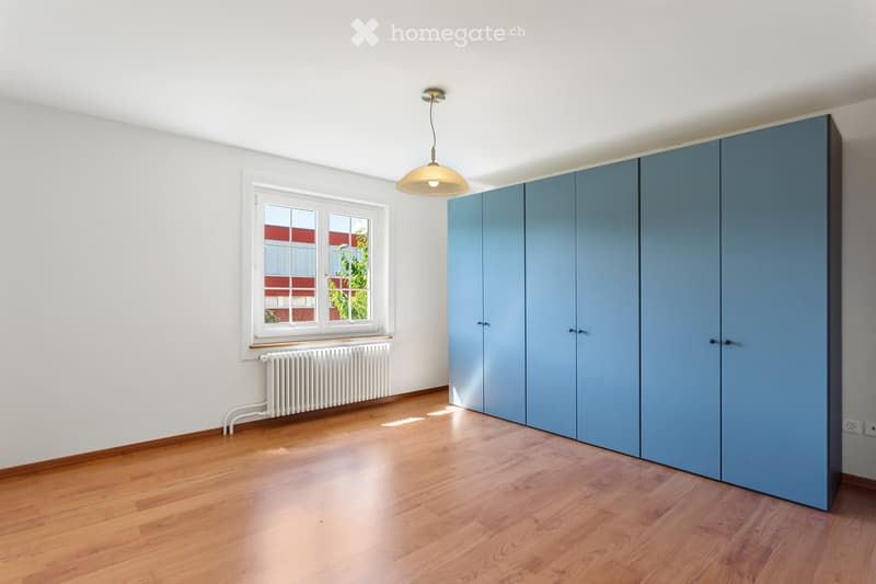 Empty room with wooden flooring, white walls, a large window with a radiator below, and a tall blue wardrobe unit.