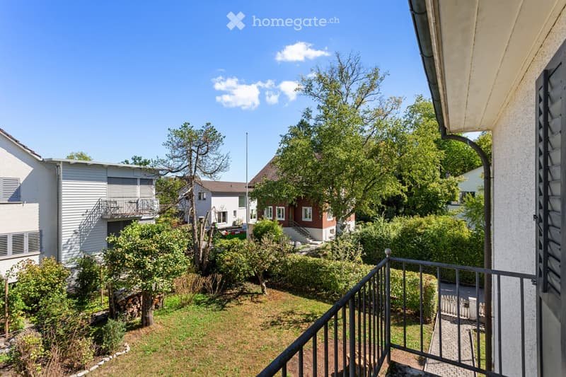 Balcony overlooking a garden area with trees, shrubs, and neighboring houses in the background.
