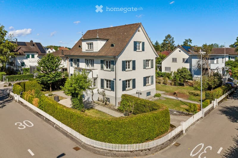 A two-story white house with blue shutters, surrounded by a well-maintained garden and white picket fence. The house features multiple windows and a pitched roof with dormer windows.