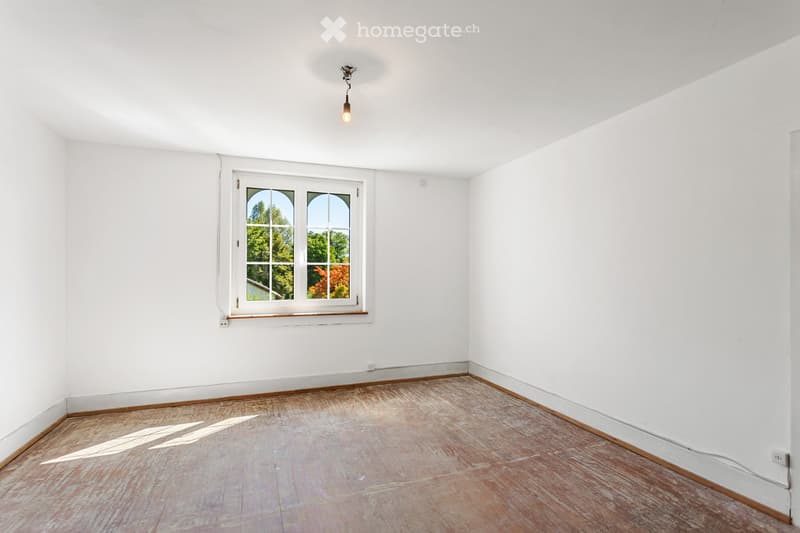 Empty room with white walls, wooden floor, and a single window with white frame and multiple panes. Natural light streams in, casting shadows on the floor.