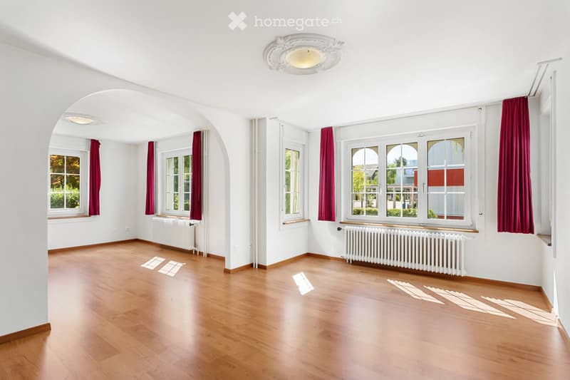 Empty room with wooden flooring, white walls, large windows with red curtains, radiators, and a decorative ceiling light fixture.
